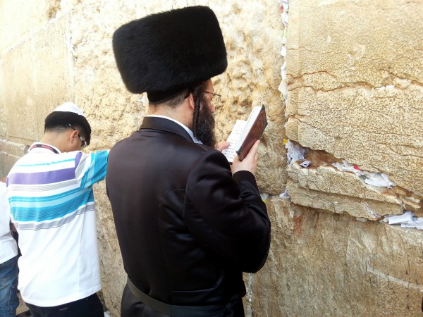 An ultra-Orthodox Jewish man and a tourist pray at the Western (Wailing) Wall in Jerusalem. The spaces between the stones in the Wall are stuffed with prayer requests. Kotel-prayer-Orthodox-tourist
